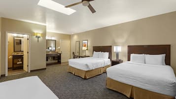 A hotel room with two beds, a ceiling fan, a desk, a chair, a mirror, and a bathroom with a sink and mirror.