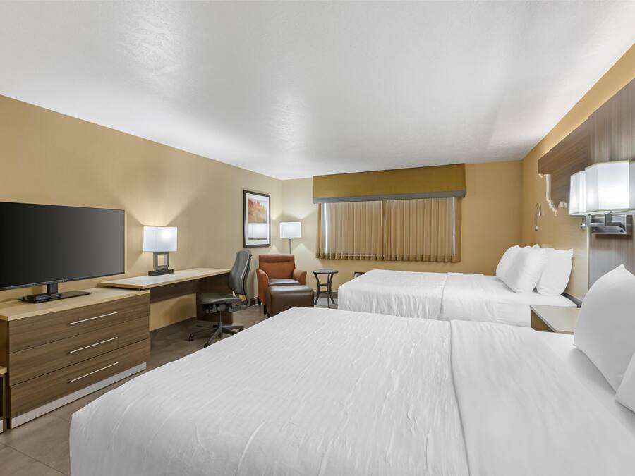 A hotel room with two beds, a desk with a flat-screen TV, a chair, a small table, and a window with blinds.