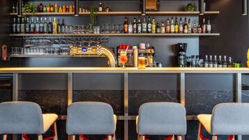 Zona de bar con estantes llenos de botellas y vasos, un grifo y una cafetera. Hay tres taburetes con detalles en rojo.