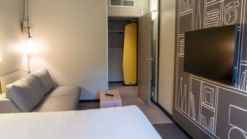 Una habitación de hotel con cama, lámpara de noche, televisor y un armario con un objeto amarillo.