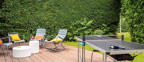 An outdoor ping pong table on a wooden deck with chairs and a table, surrounded by a lush green hedge.