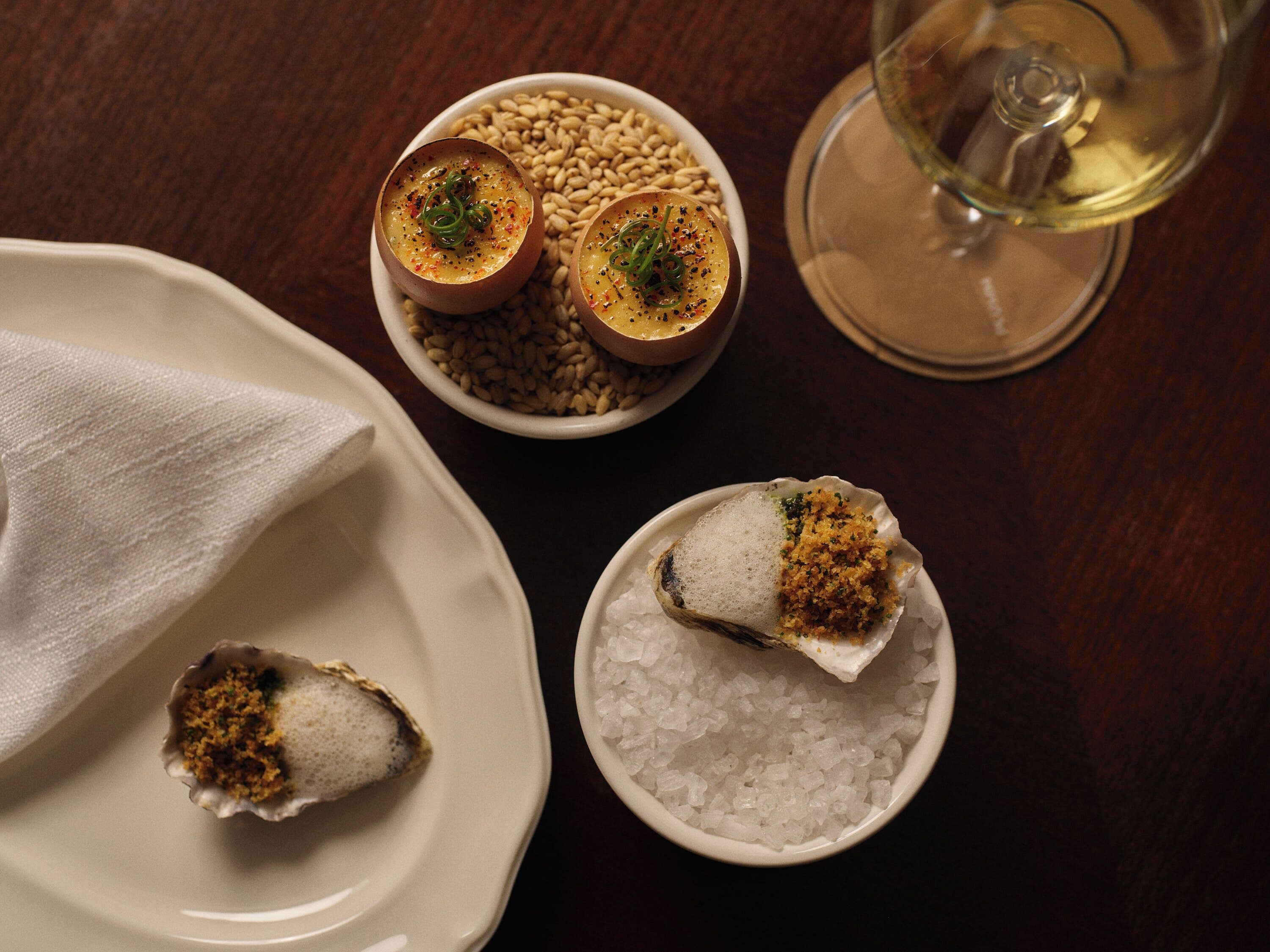 A table with a bowl of seeds, a bowl of salt, and a glass of white wine.