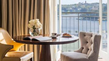 A room with a view of a body of water, a round table with a book, a vase of flowers, a chair, and a plate with food.