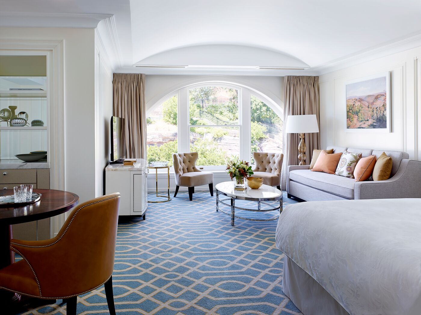 A hotel room with a large bed, a sofa, a dining table with chairs, a small round coffee table, and a view of the outdoors through a large window.
