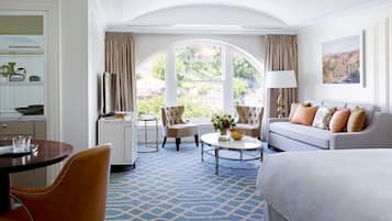 A hotel room with a large bed, a sofa, a dining table with chairs, a small round coffee table, and a view of the outdoors through a large window.