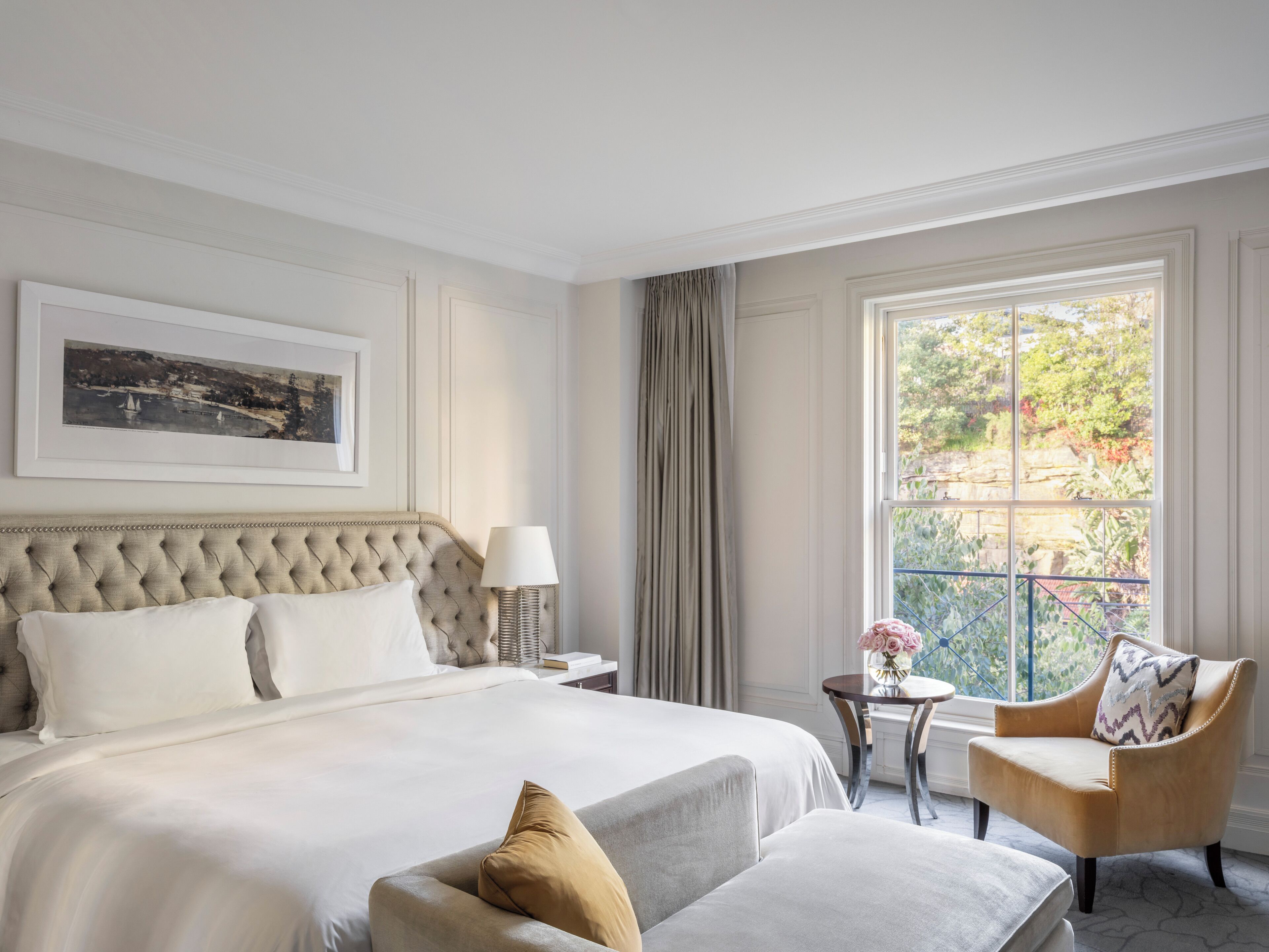 A hotel room with a large bed, a chair, a small table with a vase of flowers, and a window with a view of greenery.