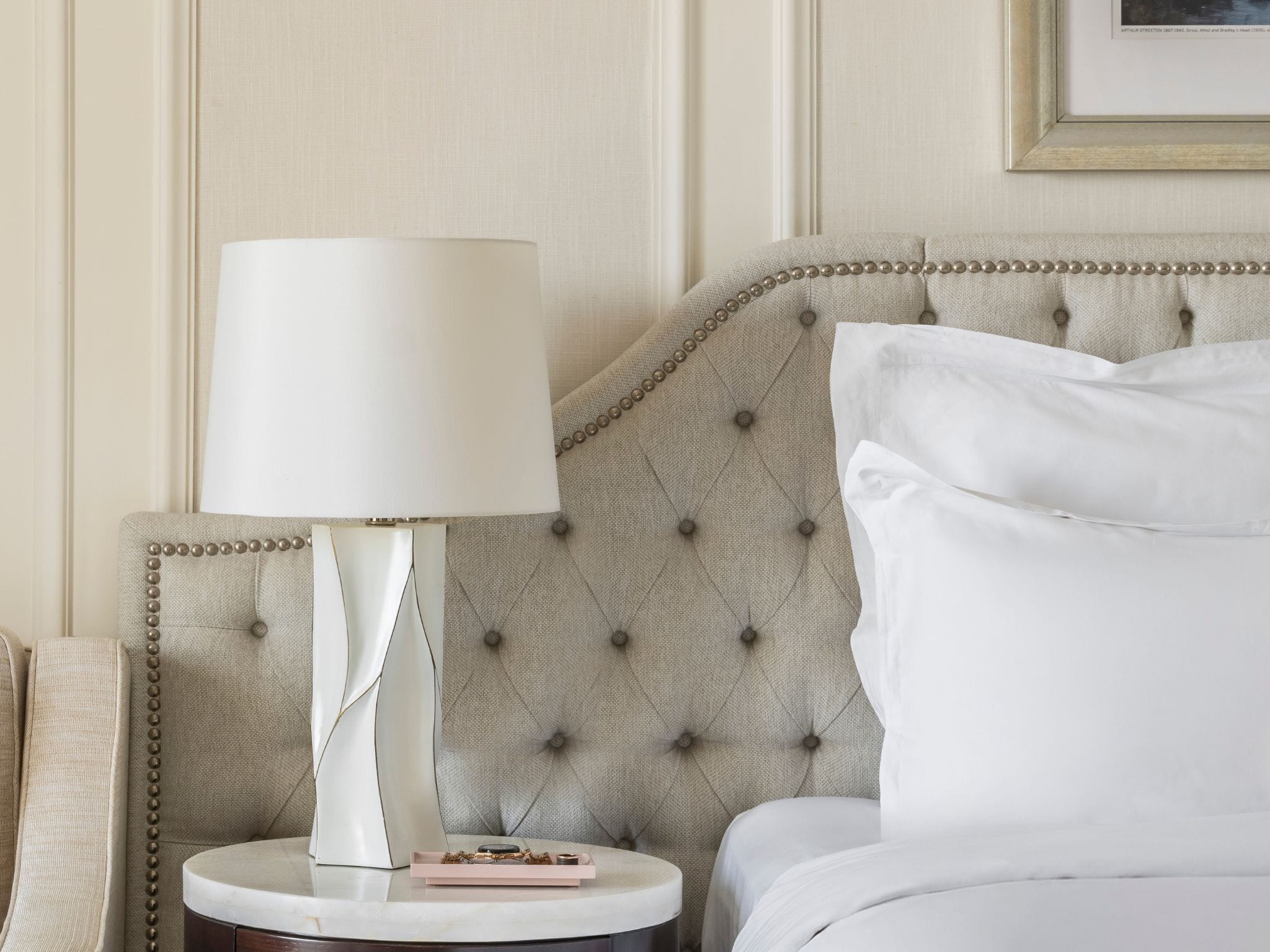 A neatly made bed with white linens, a tufted headboard, a bedside table with a lamp, and a framed picture on the wall.