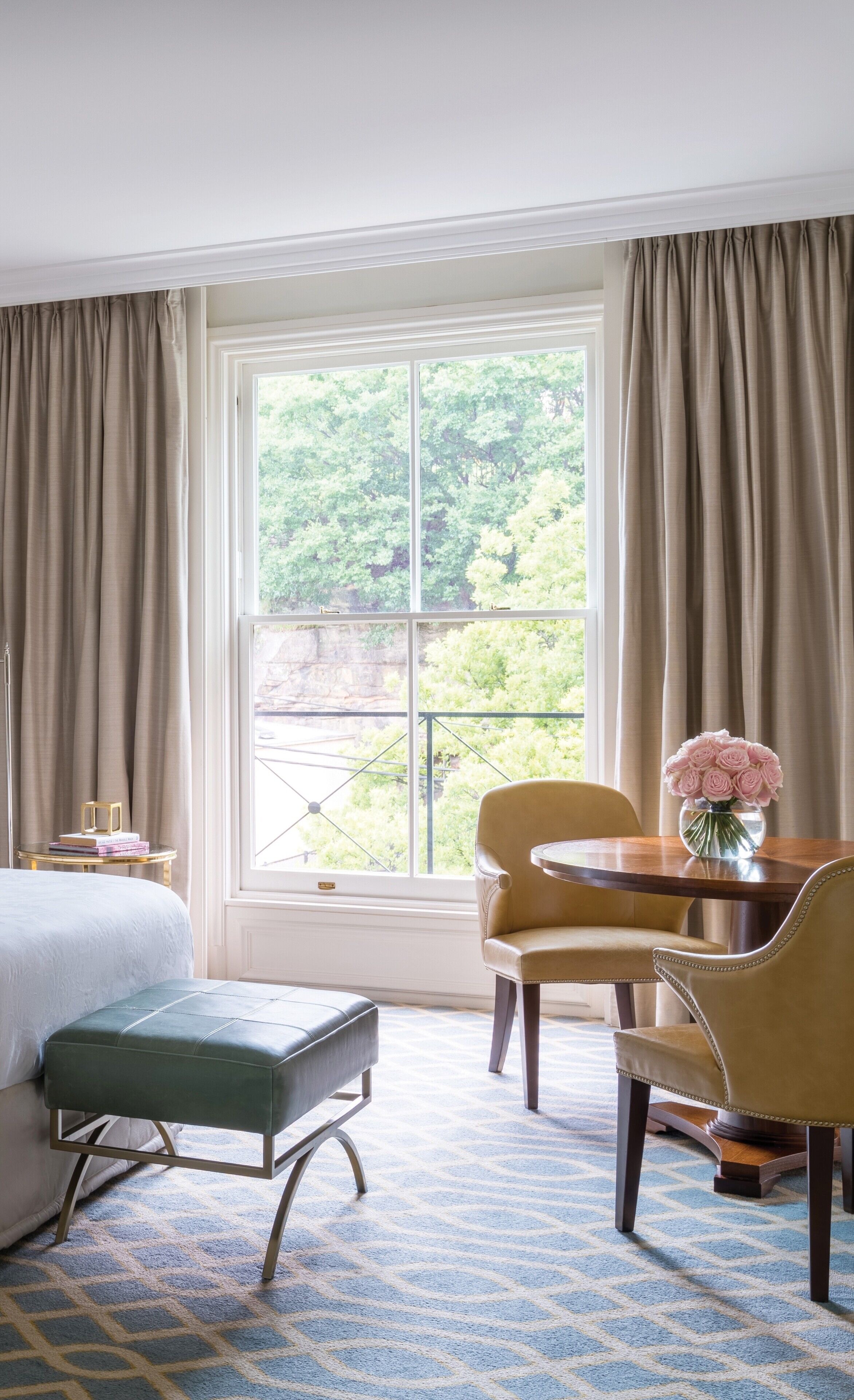 A hotel room with a bed, a small table with a vase of flowers, a chair, and a window with curtains.