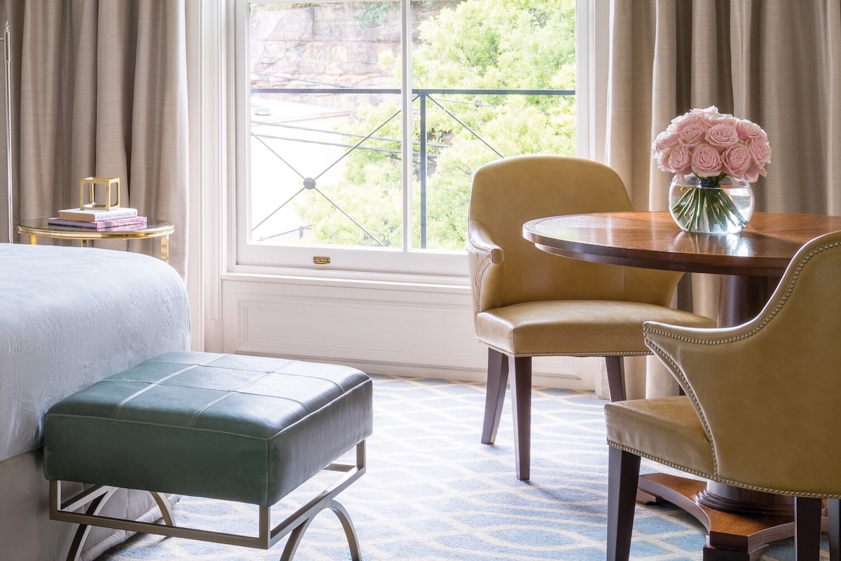 A hotel room with a bed, a small table with a vase of flowers, a chair, and a window with curtains.