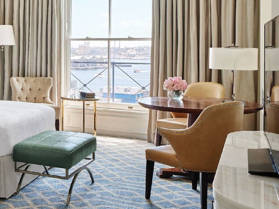 Una habitación de hotel con una cama, un reposapiés verde, una mesa redonda con un florero, una silla, un escritorio con una lámpara y un ventanal con vistas a un puente y al paisaje urbano.