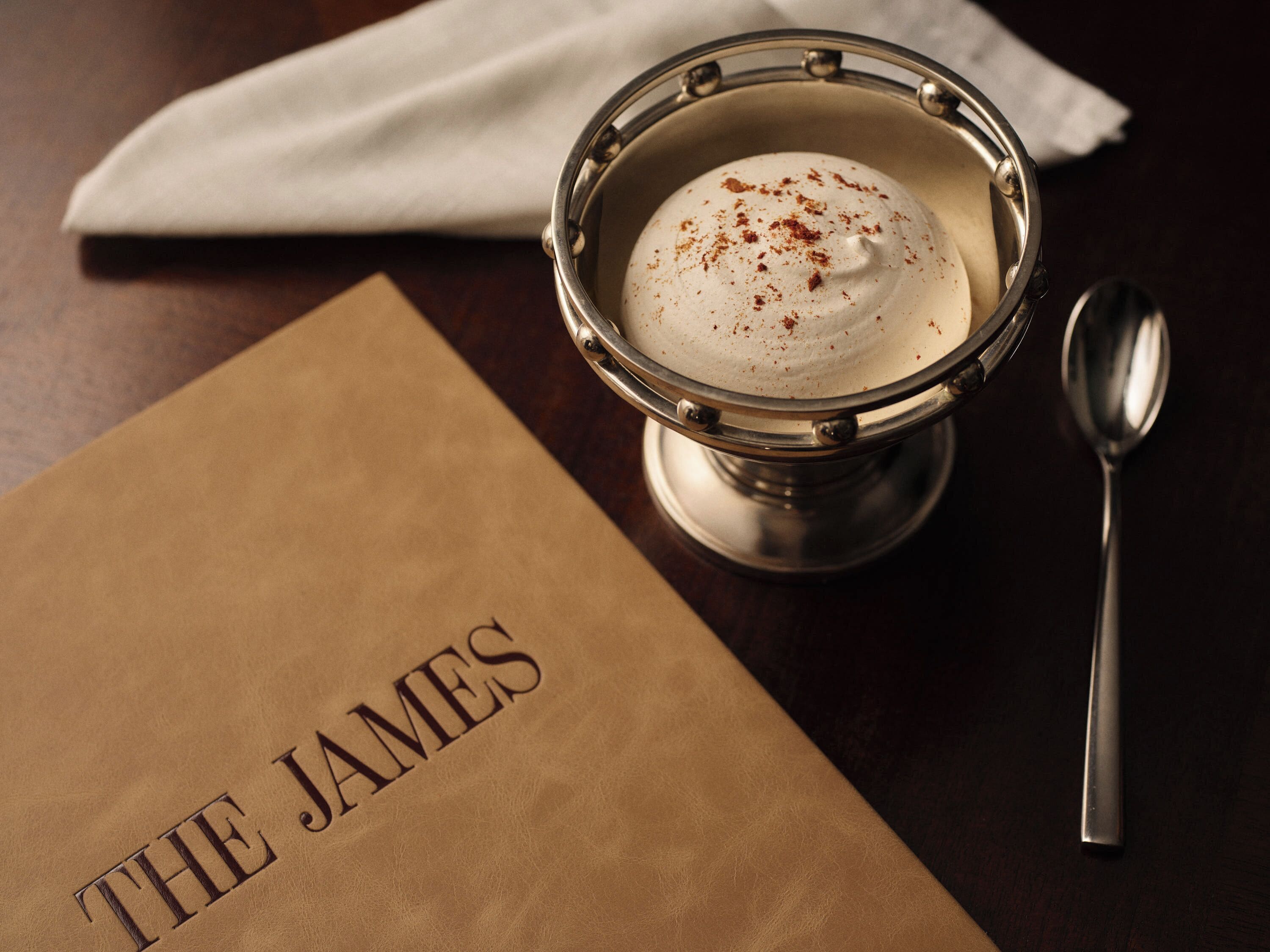 A dessert in a metal cup with a spoon beside it, on a table with a napkin and a brown paper bag with 'THE JAMES' text.