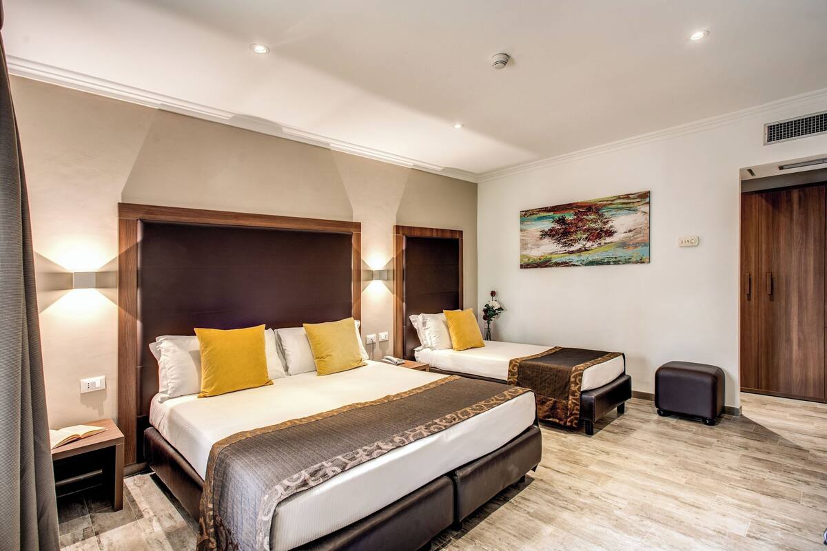 A hotel room with two beds, a small table, a bedside lamp, a painting on the wall, and a wooden door.