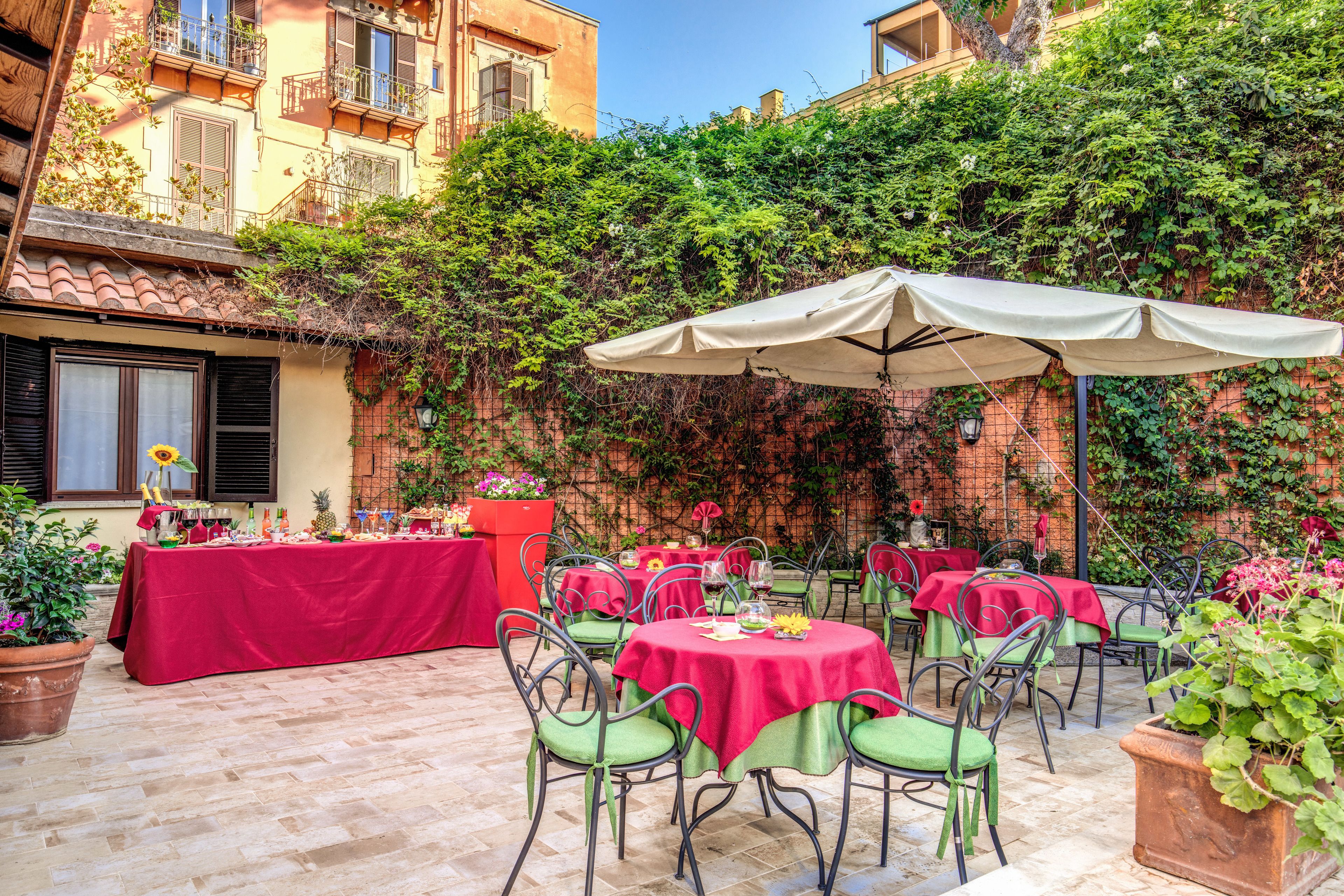 An outdoor patio with tables and chairs, covered by umbrellas, surrounded by greenery and buildings.