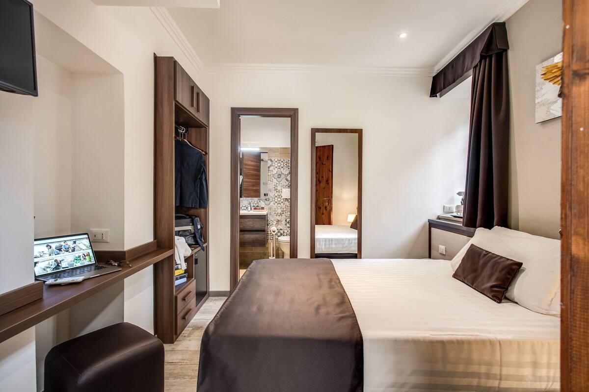 A hotel room with a bed, a desk with a laptop, a wardrobe, and a bathroom visible through an open door.