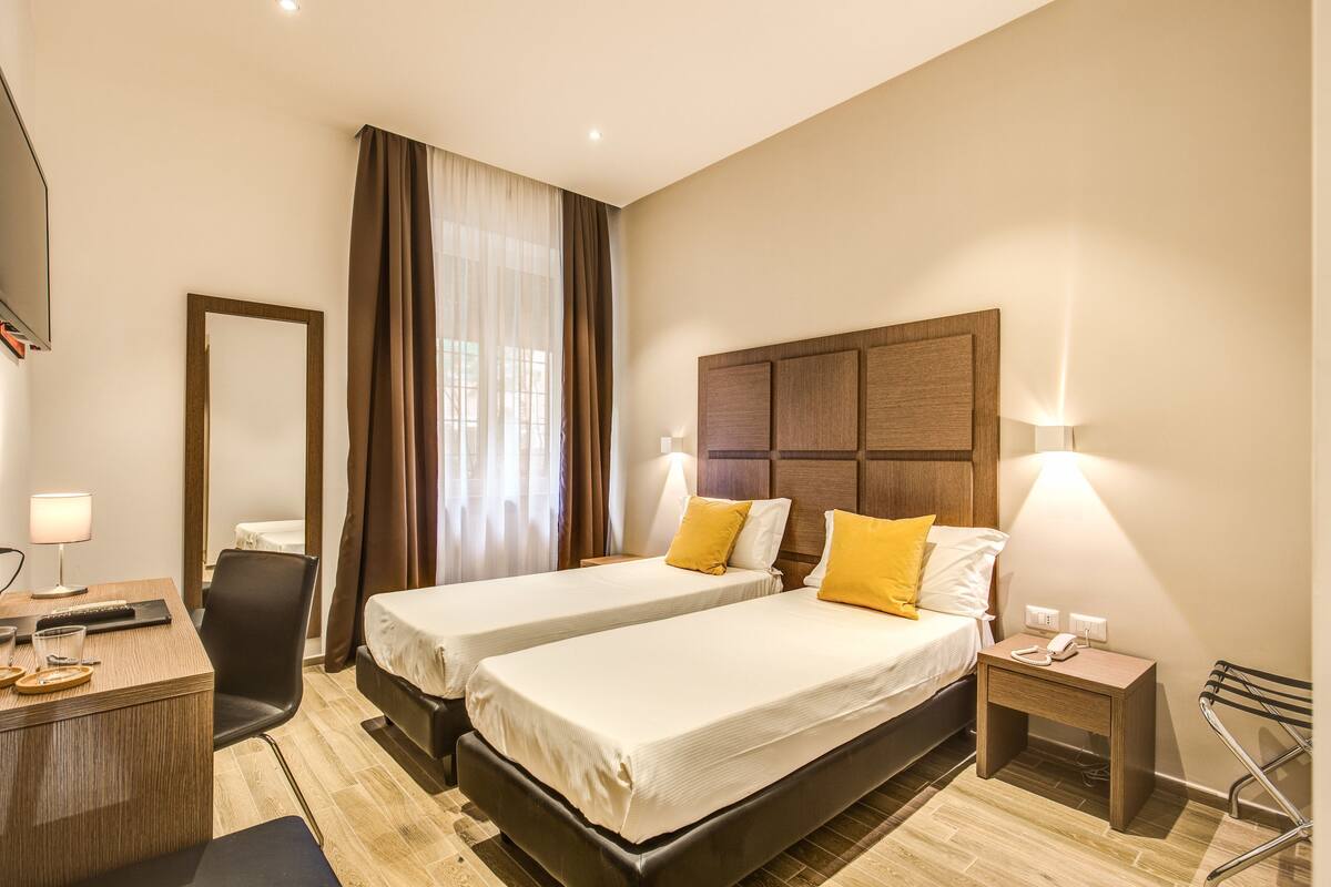 A hotel room with two beds, a desk, a chair, a TV, and a nightstand with a lamp.