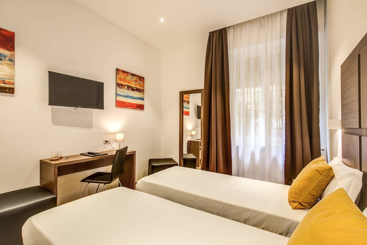 A hotel room with two beds, a desk, a chair, a TV, and a large window with curtains.