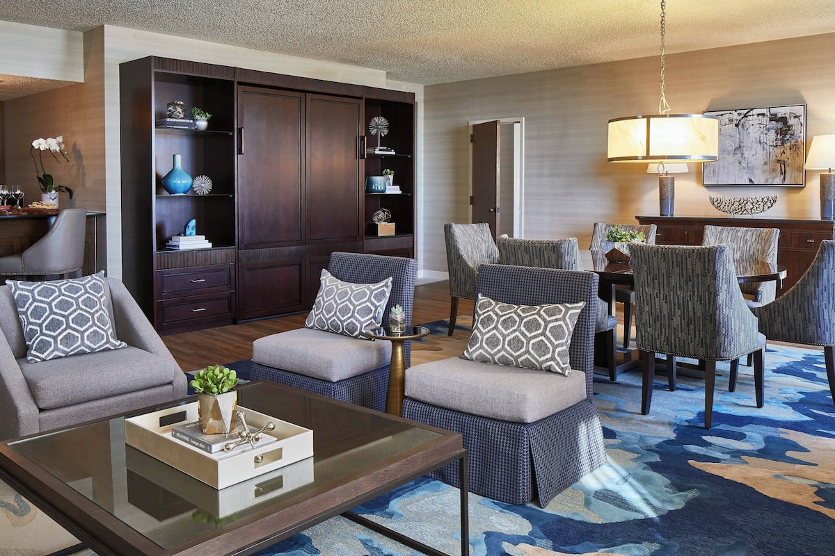 A modern living room with a glass coffee table, grey and blue upholstered chairs, a dark wooden cabinet with shelves, and a blue patterned rug.
