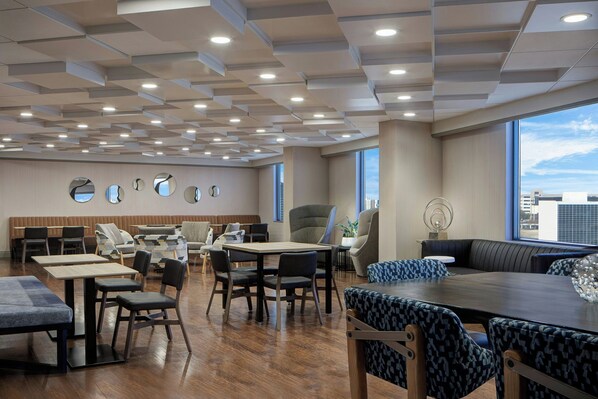 A modern hotel lobby with a wooden floor, multiple seating areas including a large round table and sofas, and a ceiling with recessed lighting.