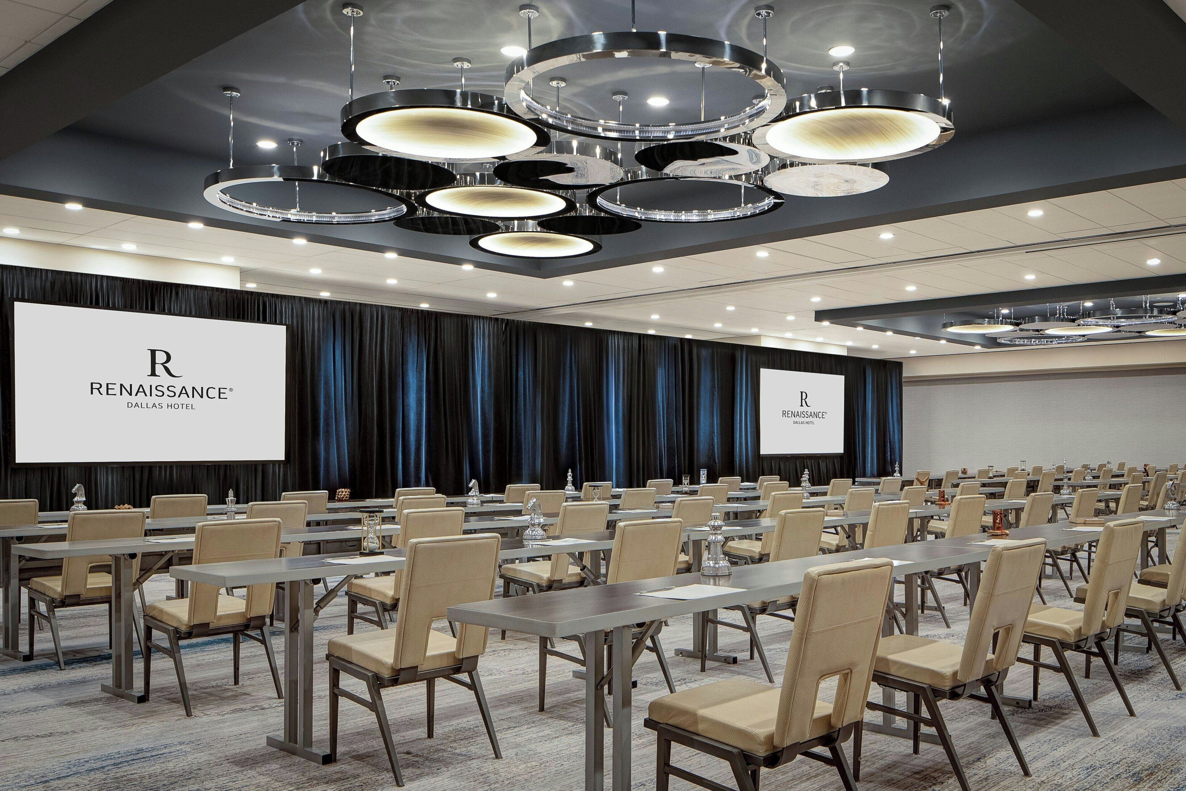 A conference room with multiple tables and chairs, a large screen displaying the hotel's name, and a modern chandelier.