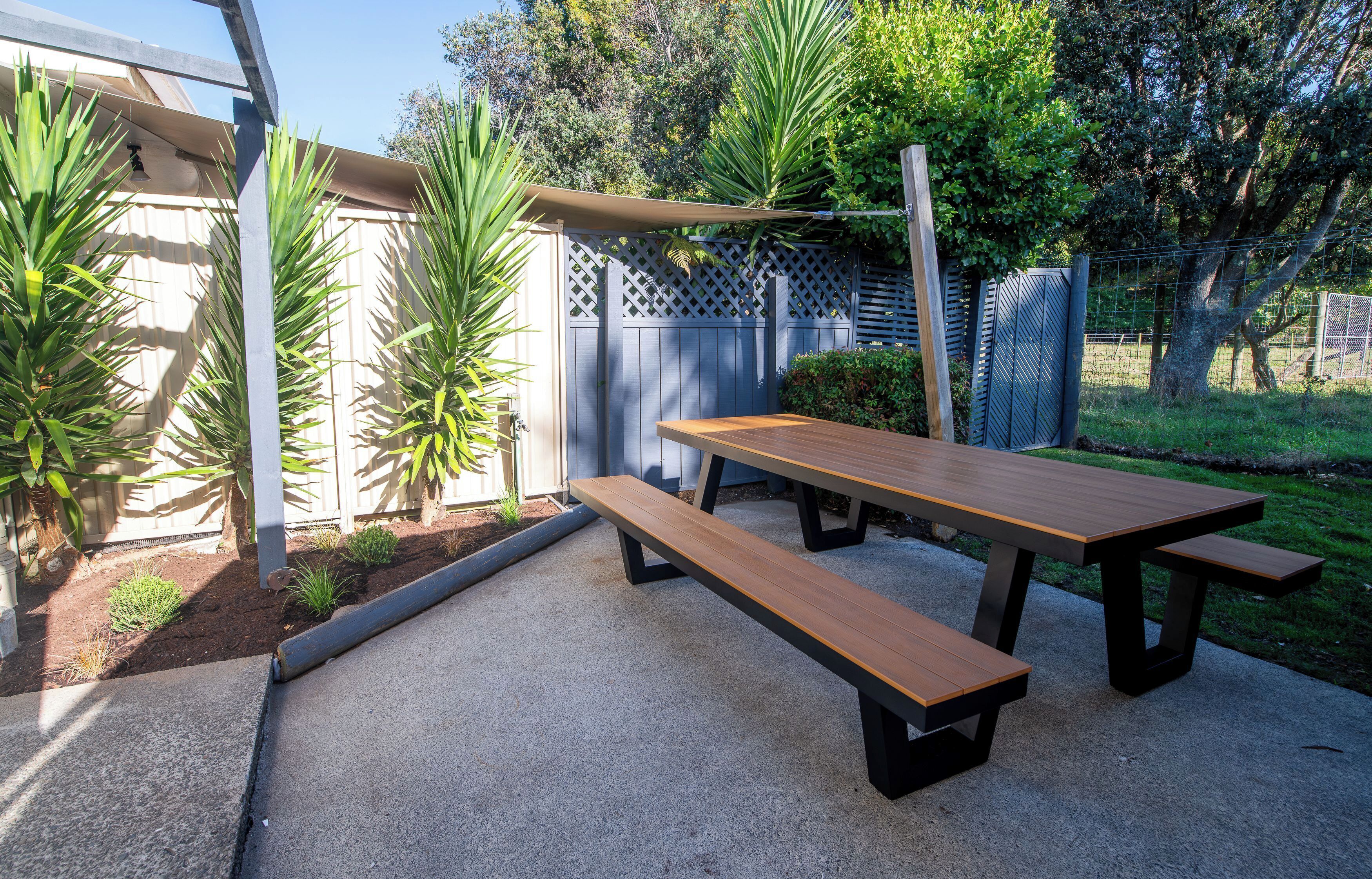 An outdoor patio area with a wooden picnic table and benches, surrounded by a fence and greenery.