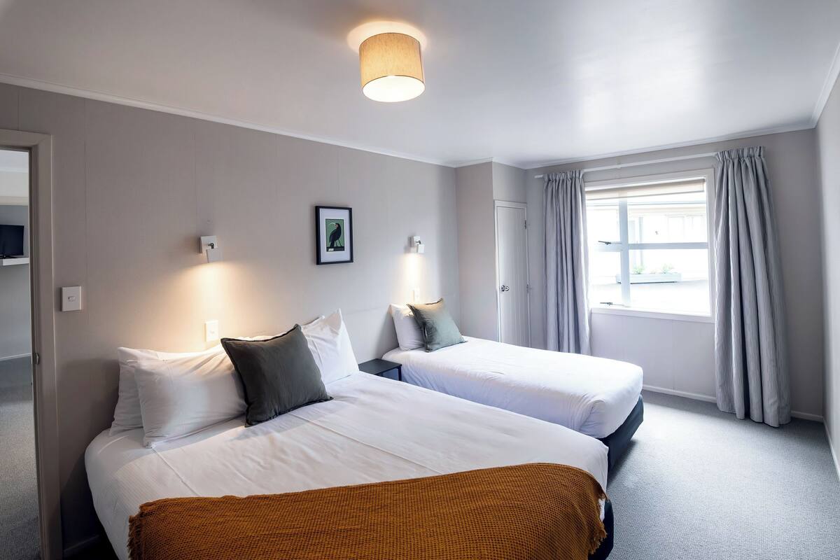 Habitación de hotel con dos camas, una ventana con cortinas, una lámpara de pared e imagen en un marco.