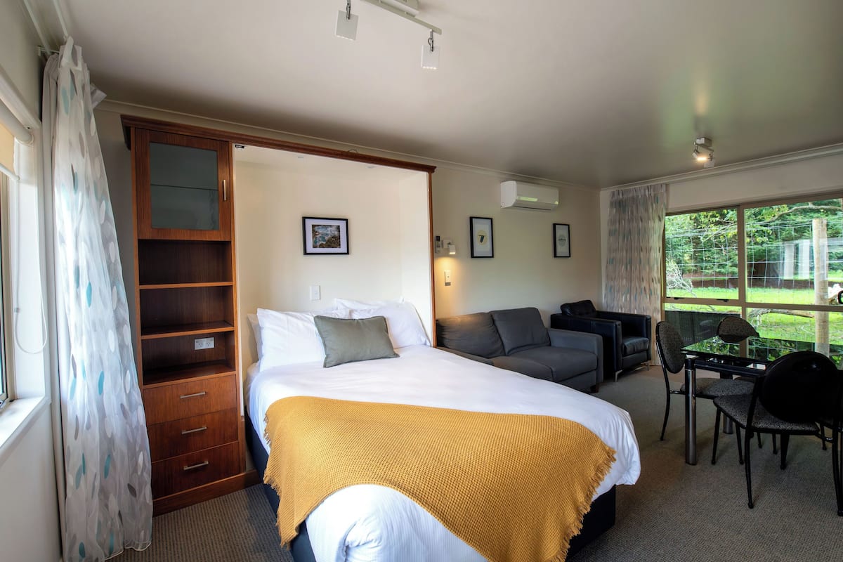 Una habitación de hotel con cama, sofá, mesa de comedor con sillas, armario de madera y un amplio ventanal con vistas al exterior.