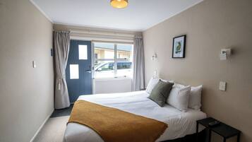 A hotel room with a bed, a bedside table with a remote control, a window with curtains, and a wall-mounted light fixture.