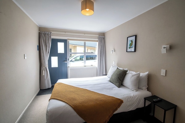 A hotel room with a bed, a bedside table with a remote control, a window with curtains, and a wall-mounted light fixture. - Ripple Rotorua (Rotorua)