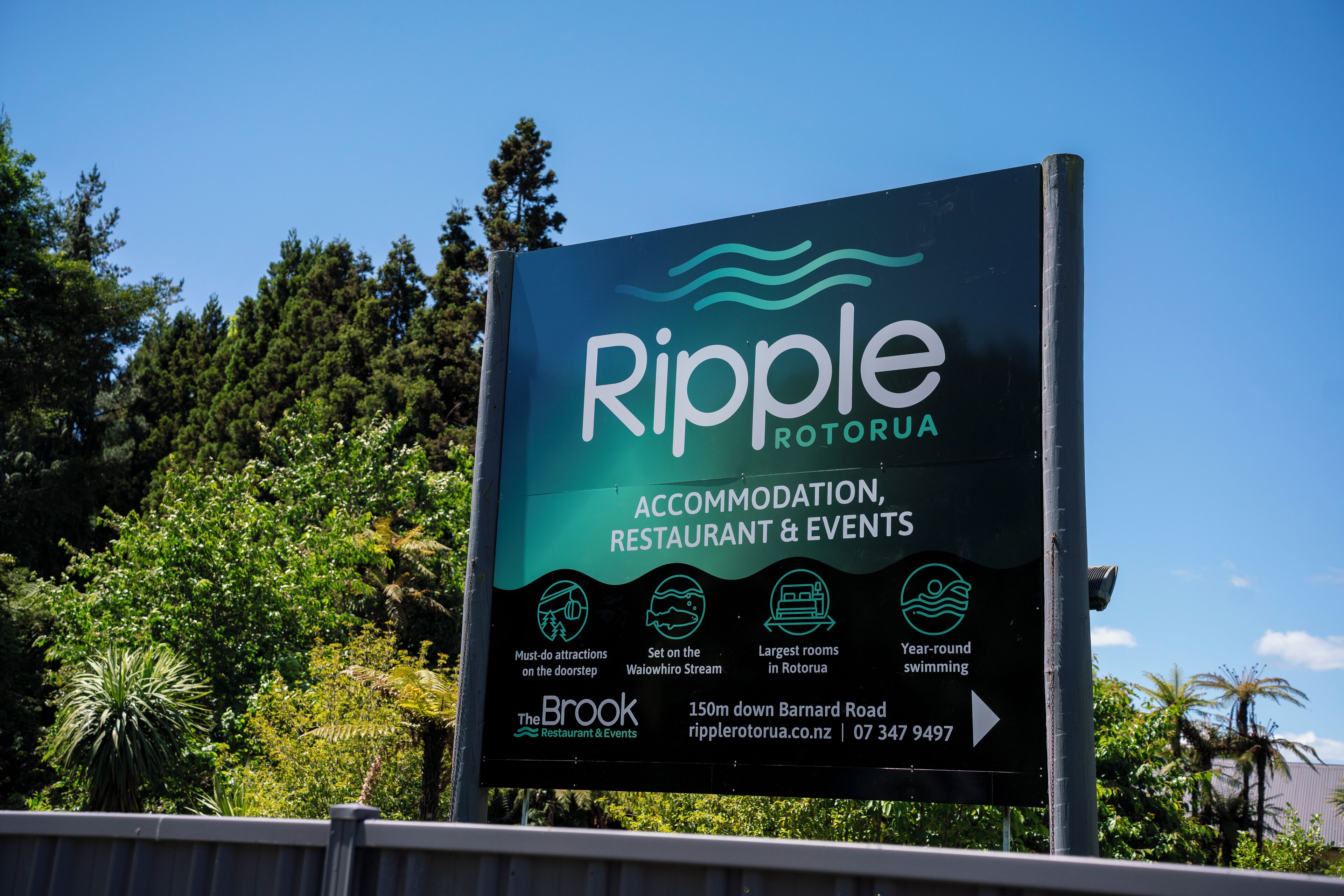 A sign for Ripple Rotoria, highlighting accommodation, restaurant, and events. It mentions must-do attractions, a restaurant on the Waikoro Stream, and year-round swimming.