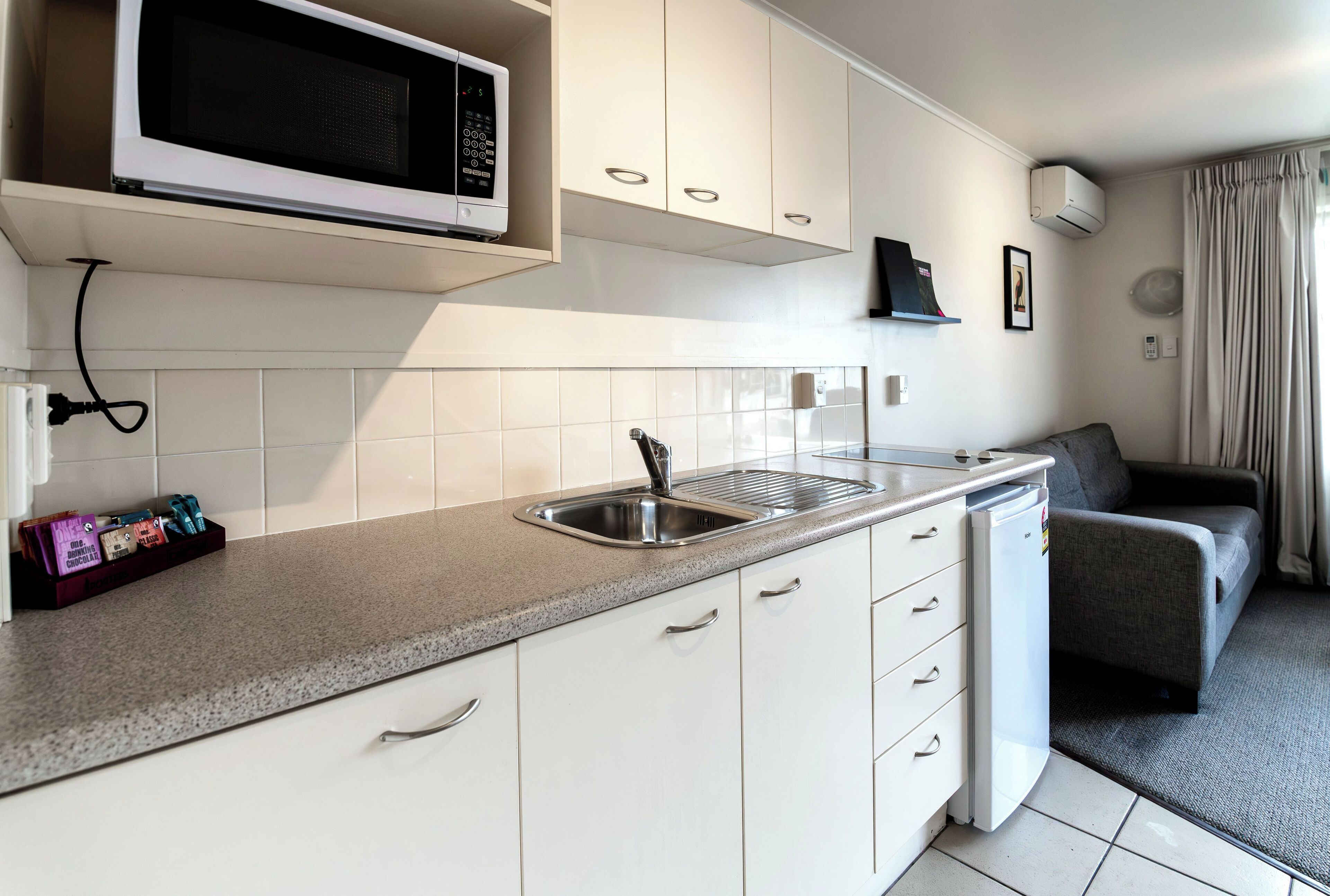 A compact kitchen with a microwave, sink, and white cabinets. There is a gray sofa and a small refrigerator in the adjacent living area.