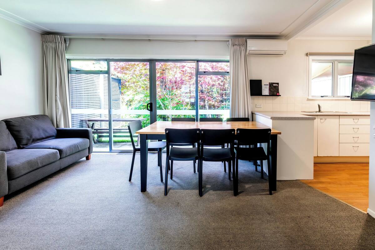 Un salón moderno con un sofá gris, una mesa de comedor de madera con cuatro sillas y un amplio ventanal corredero que da a un balcón.