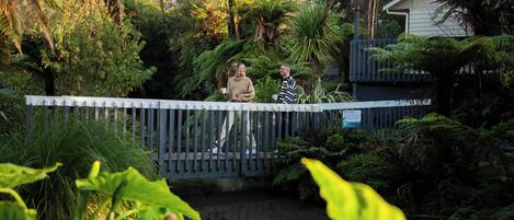 Un pont en bois enjambe un ruisseau dans un jardin luxuriant, avec un feuillage dense, et une maison se profile à l’arrière-plan.