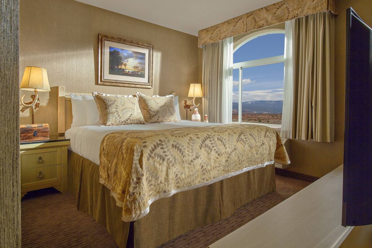 A hotel room with a large bed, two bedside tables with lamps, a painting on the wall, and a view of the mountains through the window.