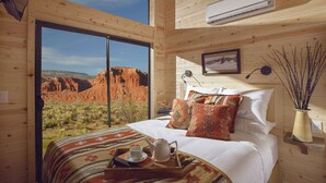 Een houten kamer met een groot bed, een raam met uitzicht op rode rotsformaties, een airconditioning en een nachtkastje met een vaas takken.