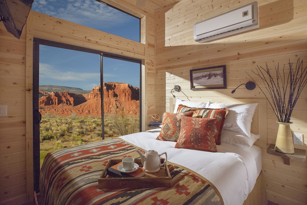 A wooden cabin room with a large bed, a window with a view of red rock formations, an air conditioning unit, and a bedside table with a vase of branches.