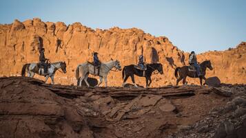 Um grupo de pessoas a cavalo em terreno rochoso, com uma grande formação rochosa avermelhada ao fundo.