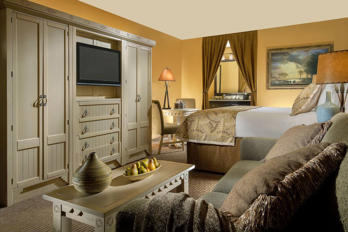 A hotel room with a bed, a television, a desk with a vase and fruit bowl, a chair, and a wardrobe.