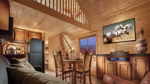 Een gezellig chaletinterieur met houten wanden, een eethoek, een woonkamer met een leren bank en een tv waarop een paardenrace te zien is.