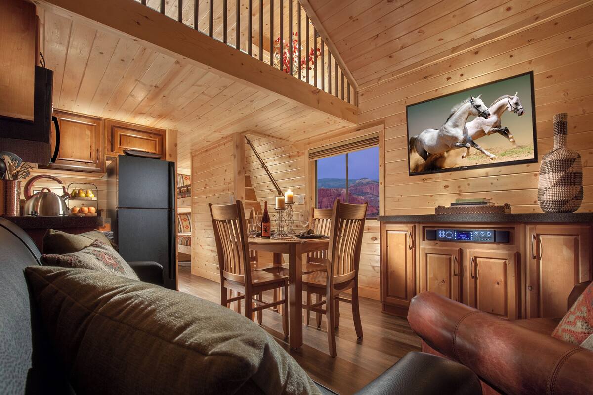 A cozy cabin interior with wooden walls, a dining area, a living room with a leather couch, and a mounted TV displaying a horse race.