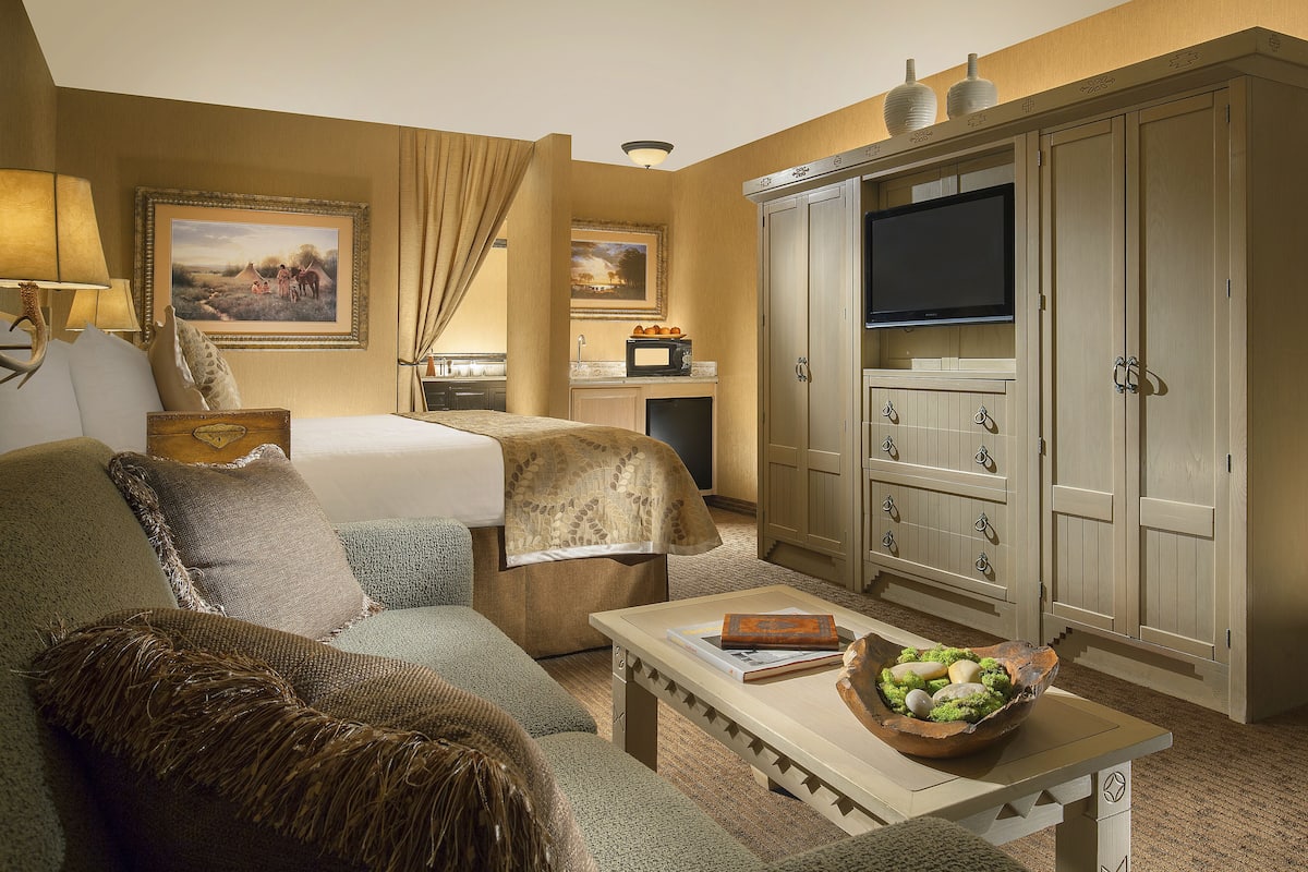 A hotel room with a bed, a sofa, a television, a desk with a bowl of fruit, and a painting on the wall.