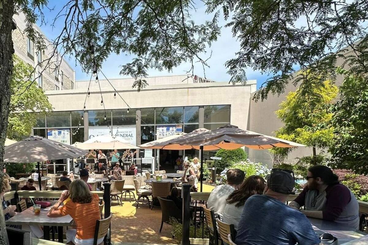 An outdoor dining area with tables and umbrellas, surrounded by trees and a building with large windows.