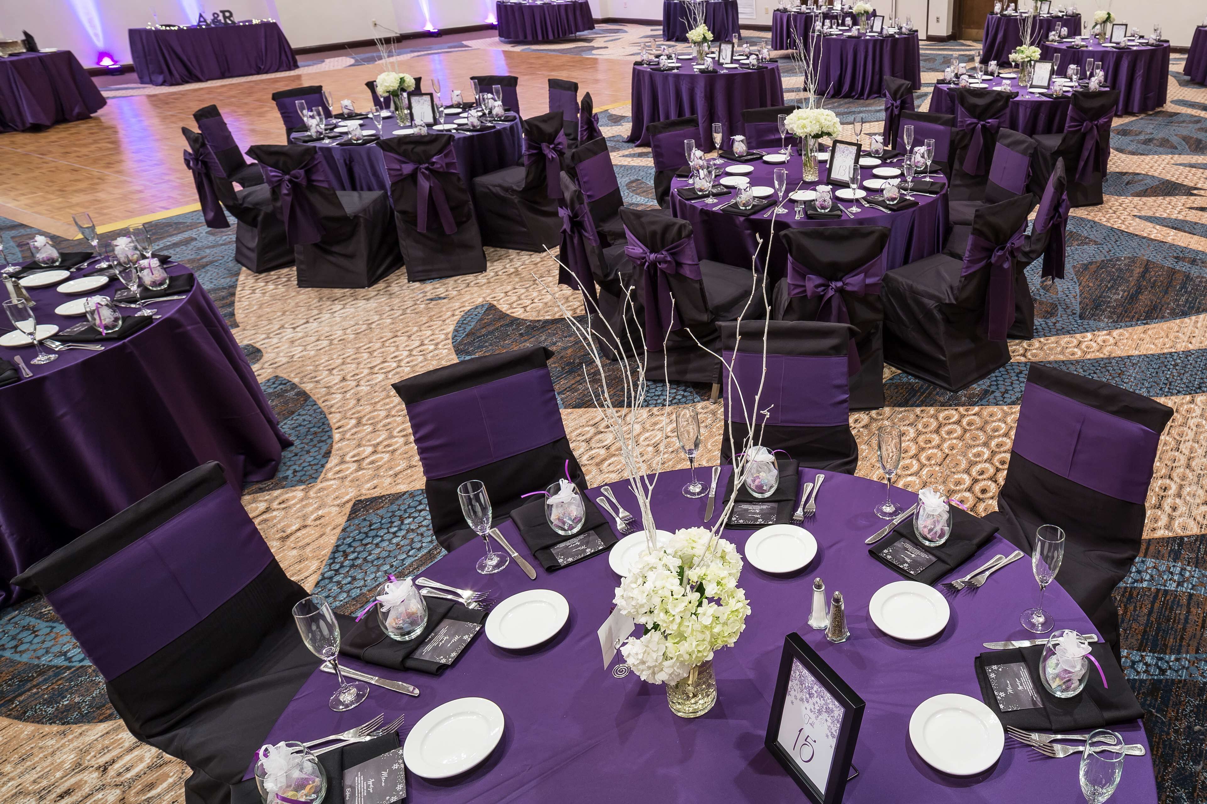 A banquet hall with round tables set for a formal event, featuring purple tablecloths and black chairs with purple sashes.