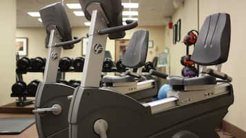 A stationary bike with a digital display, a wall-mounted mirror, and a rack of dumbbells in a gym.