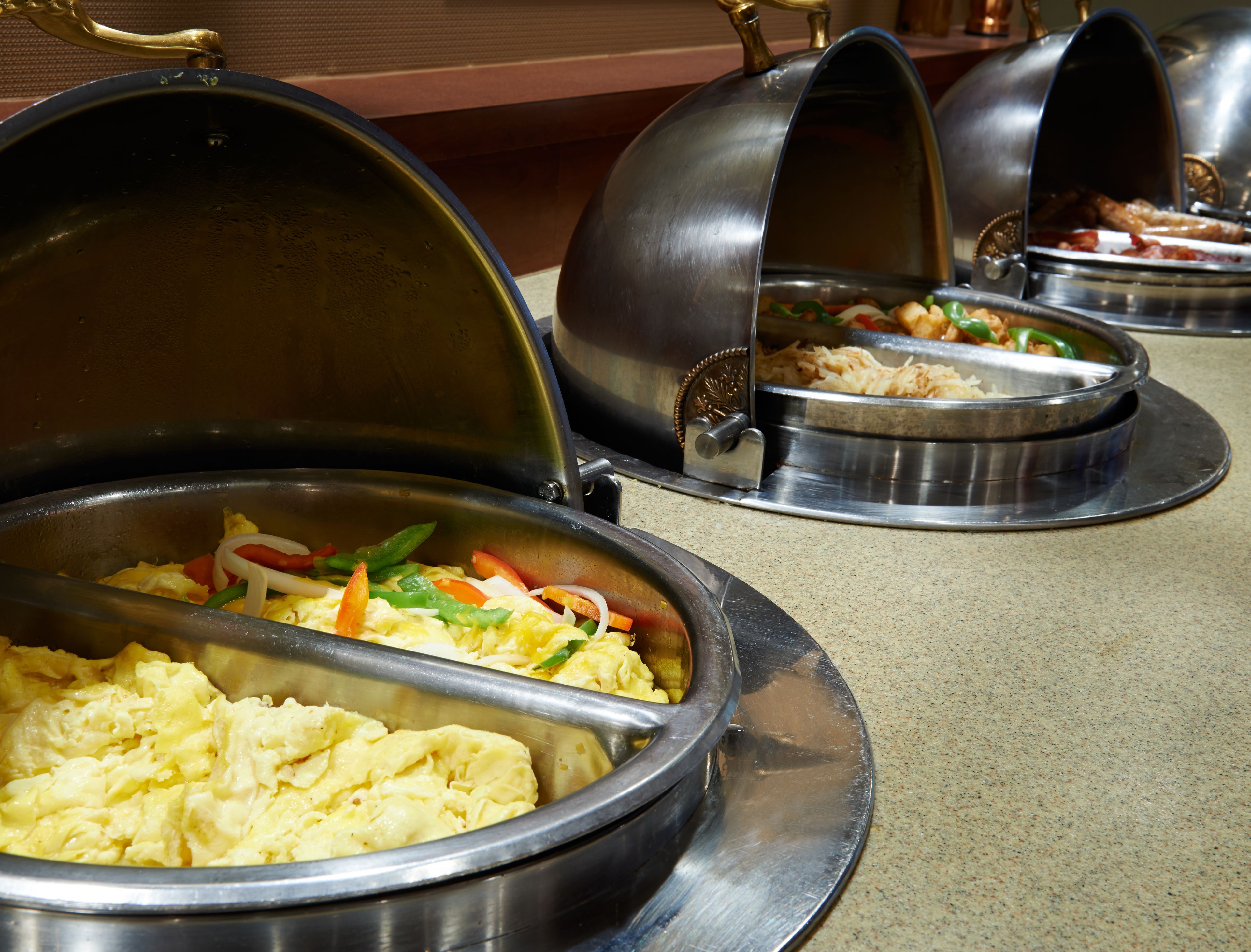 Buffet-style food service with various dishes, including scrambled eggs and vegetables, served in metal containers with lids.
