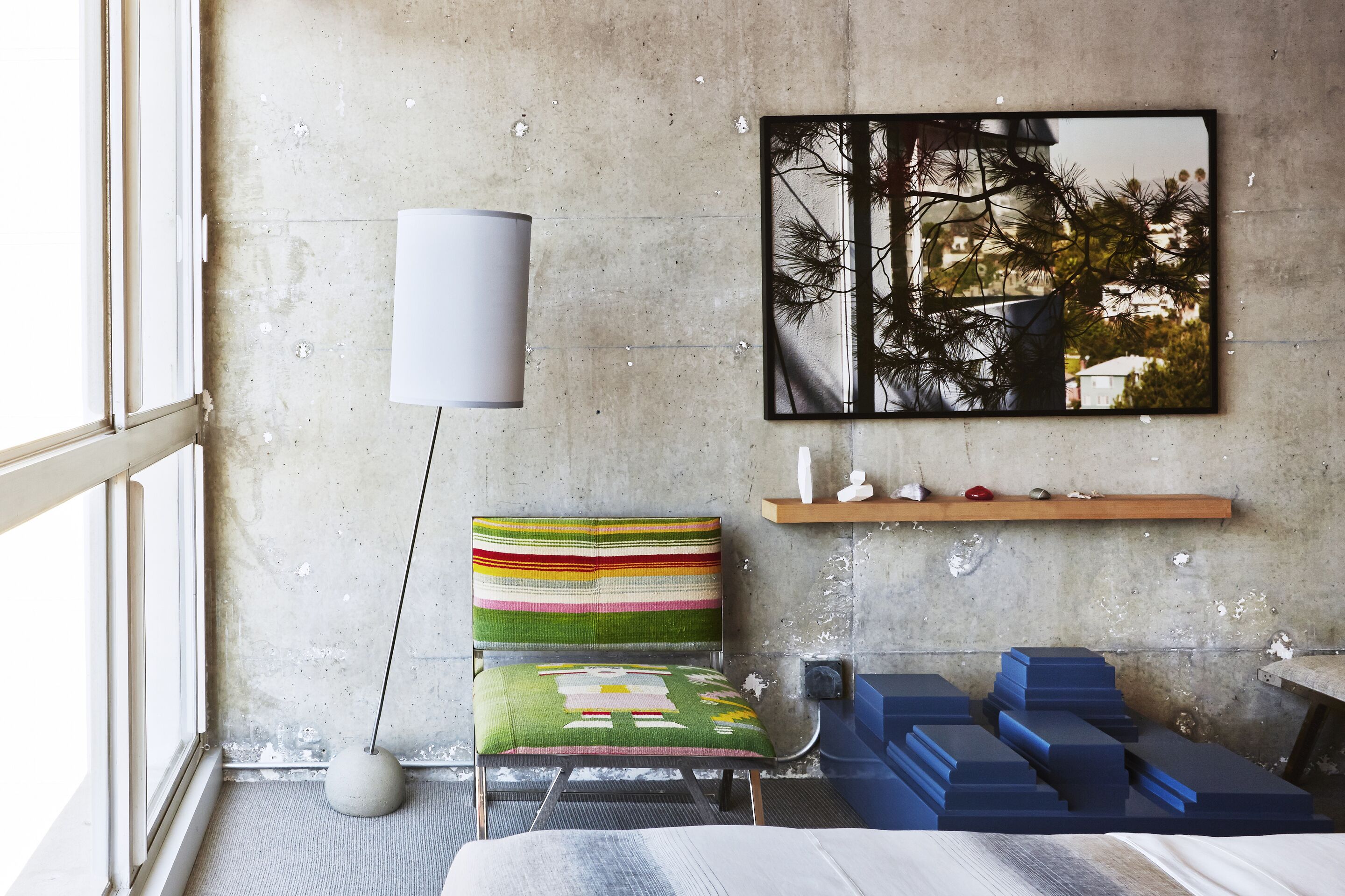 Une pièce moderne avec un mur en béton, une grande fenêtre, une chaise à rayures colorées, un lampadaire blanc, un tableau encadré et une étagère en bois ornée d’objets décoratifs.