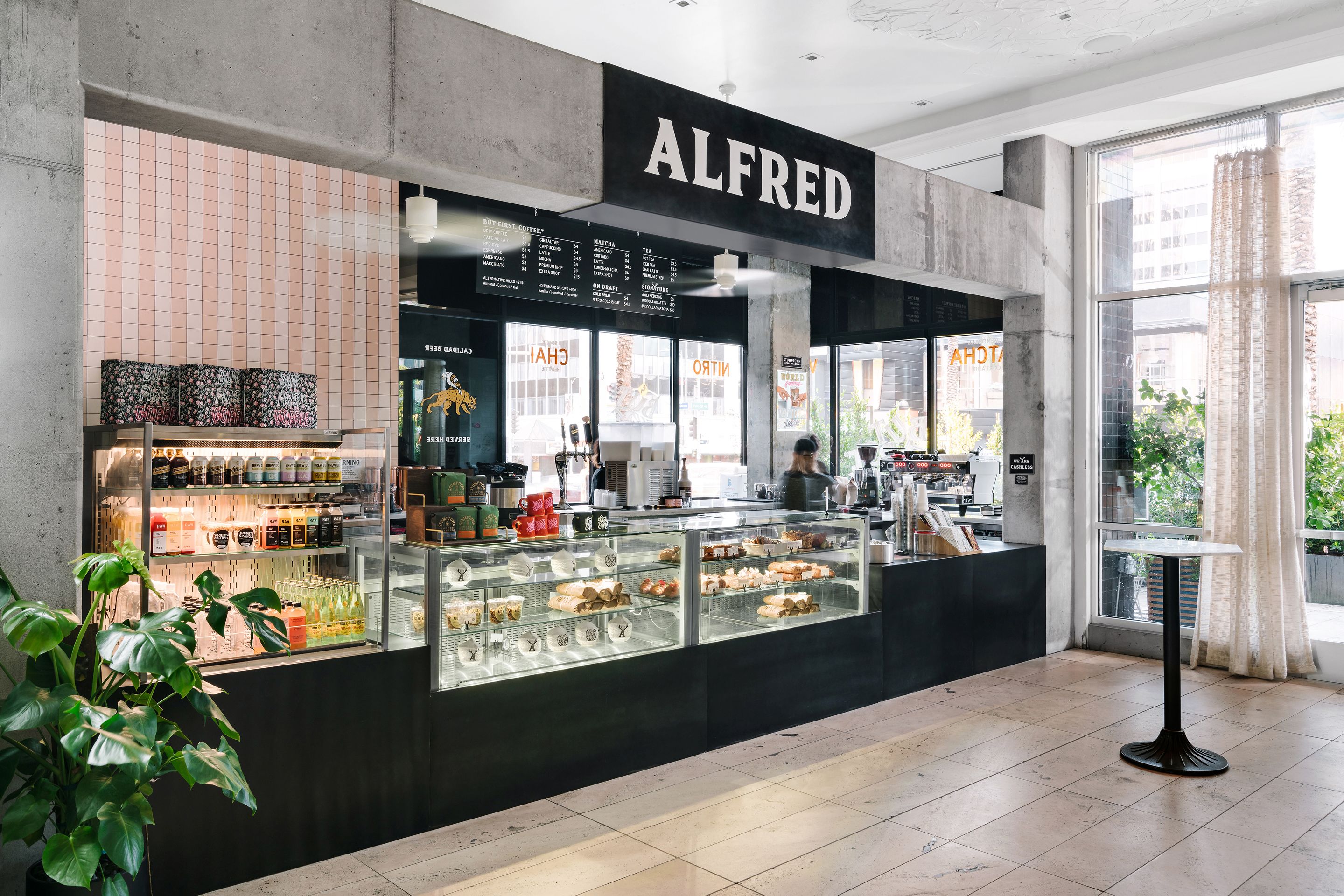 A coffee shop named 'ALFRED' with a menu board, display cases of pastries, and a seating area with a table and chairs.