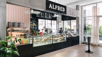 A coffee shop named 'ALFRED' with a menu board, display cases of pastries, and a seating area with a table and chairs.
