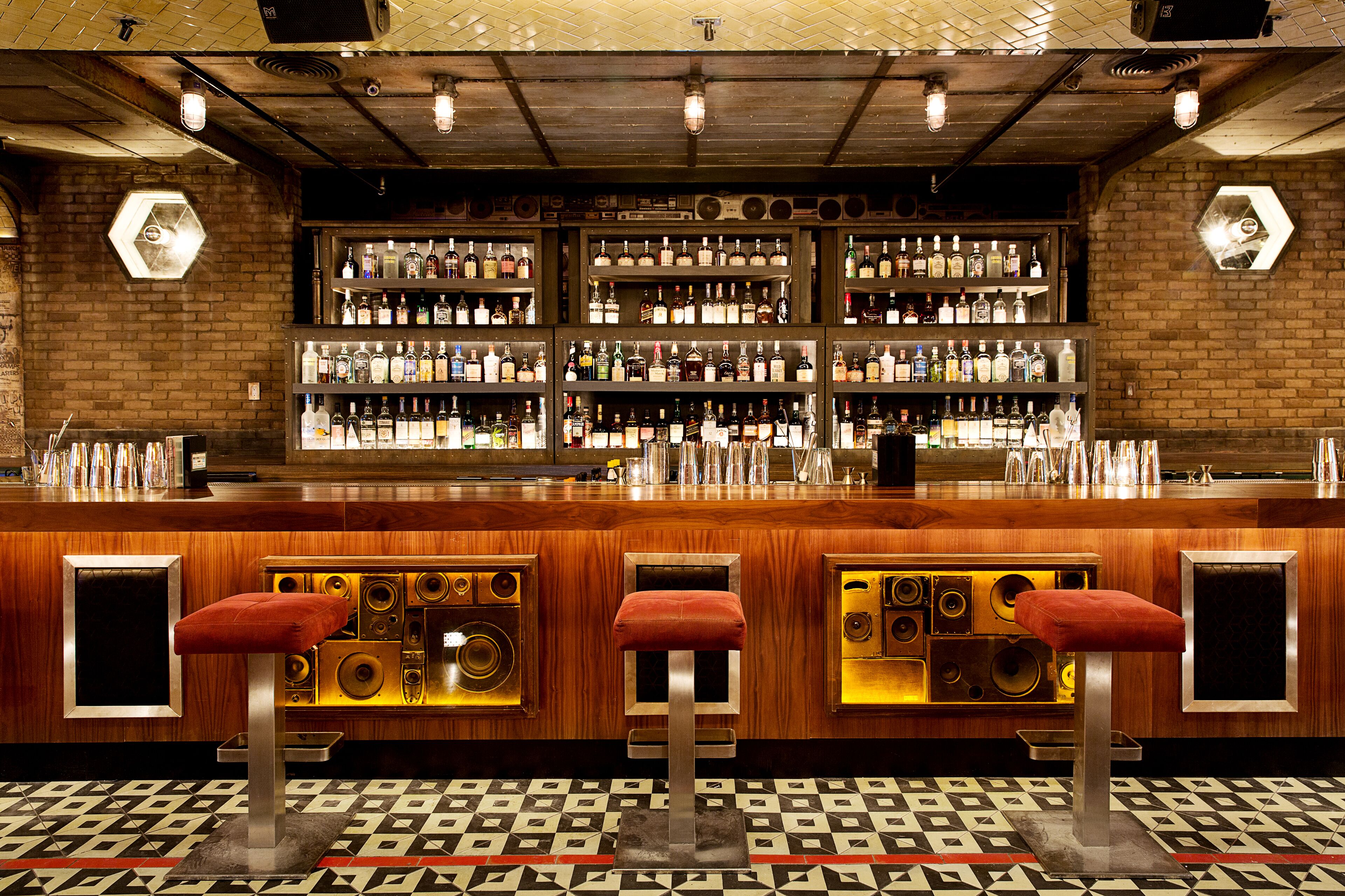 Un bar avec plusieurs étagères remplies de bouteilles, un comptoir en bois et trois tabourets dotés de coussins rouges.