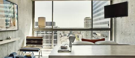 A modern hotel room with a large window, a flat-screen TV, a desk, and a comfortable seating area.