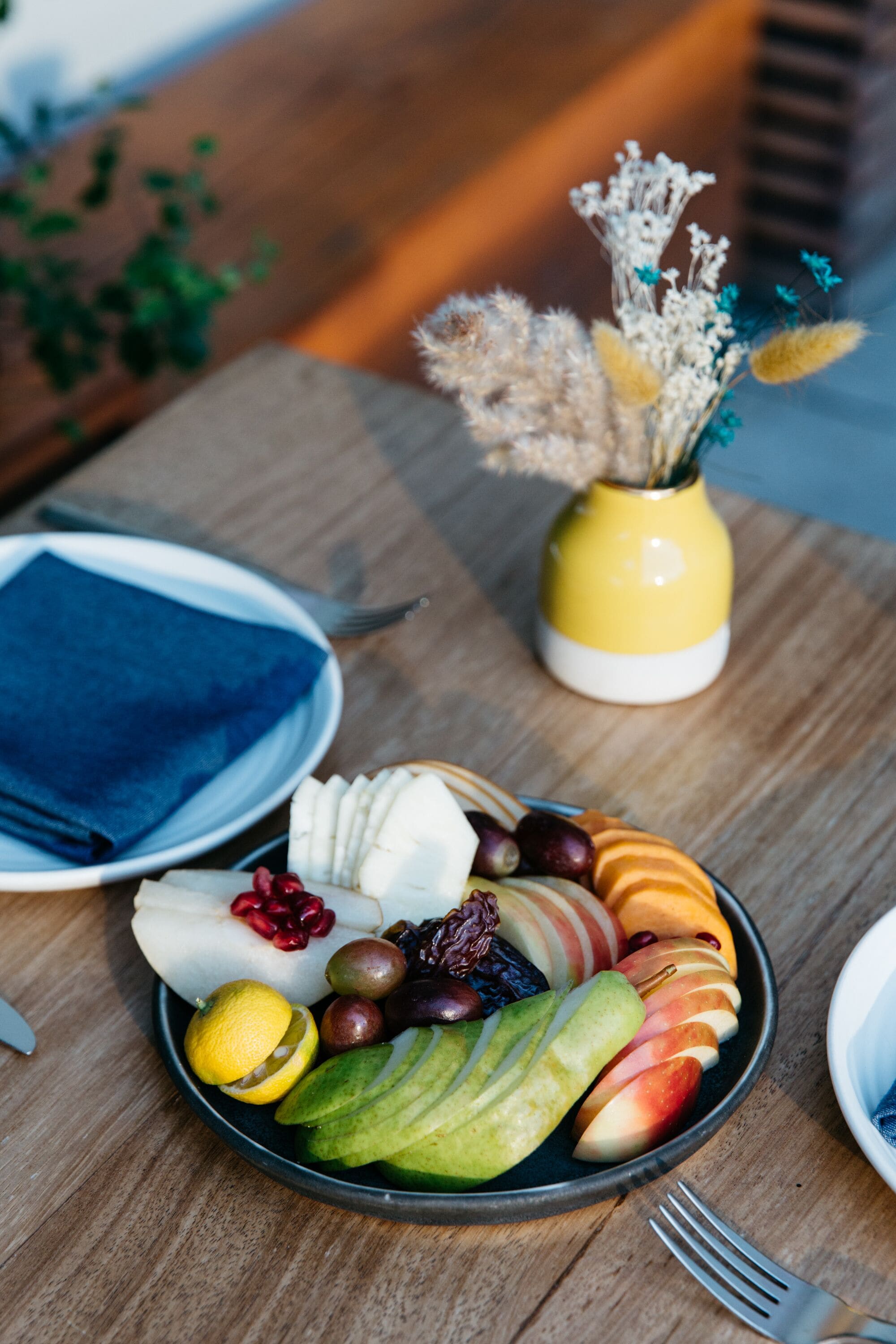 Une table en bois sur laquelle se trouve une assiette de fruits coupés et un vase jaune contenant des fleurs séchées.